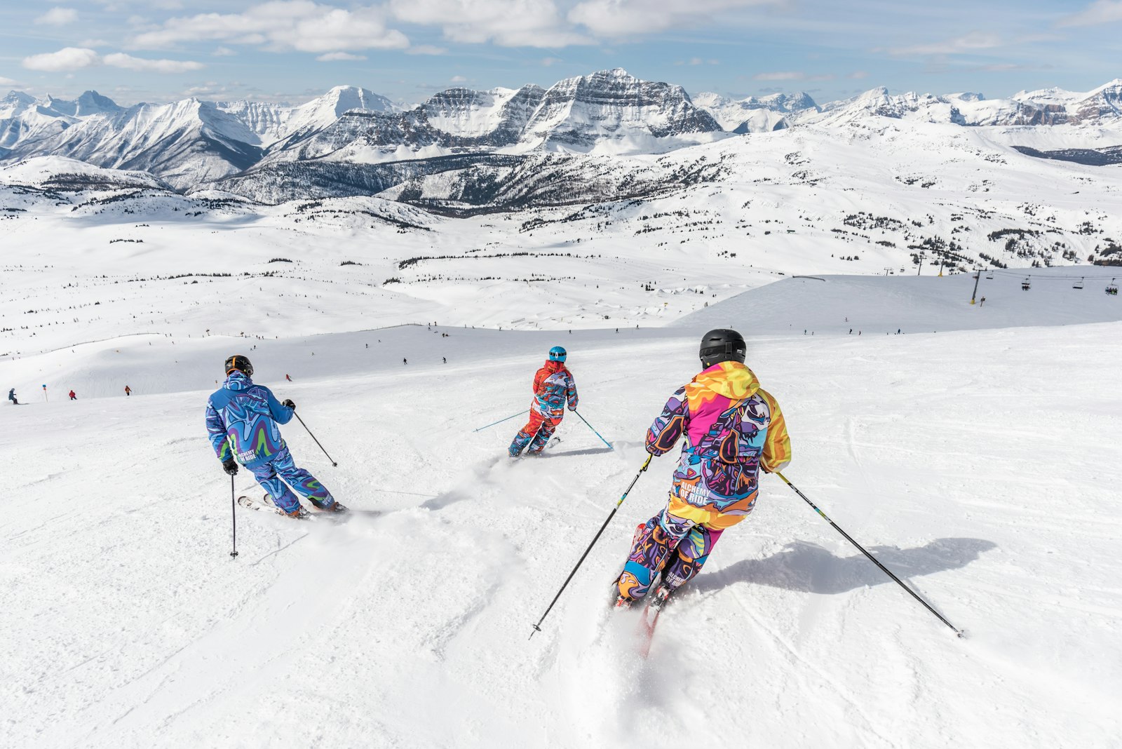 Skiers crossing a wide sunlit slope with rocky peaks behind them.